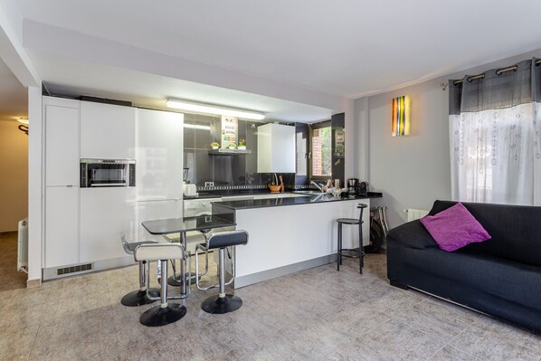 A modern kitchen with a central island, bar stools, and a black sofa with a purple throw.
