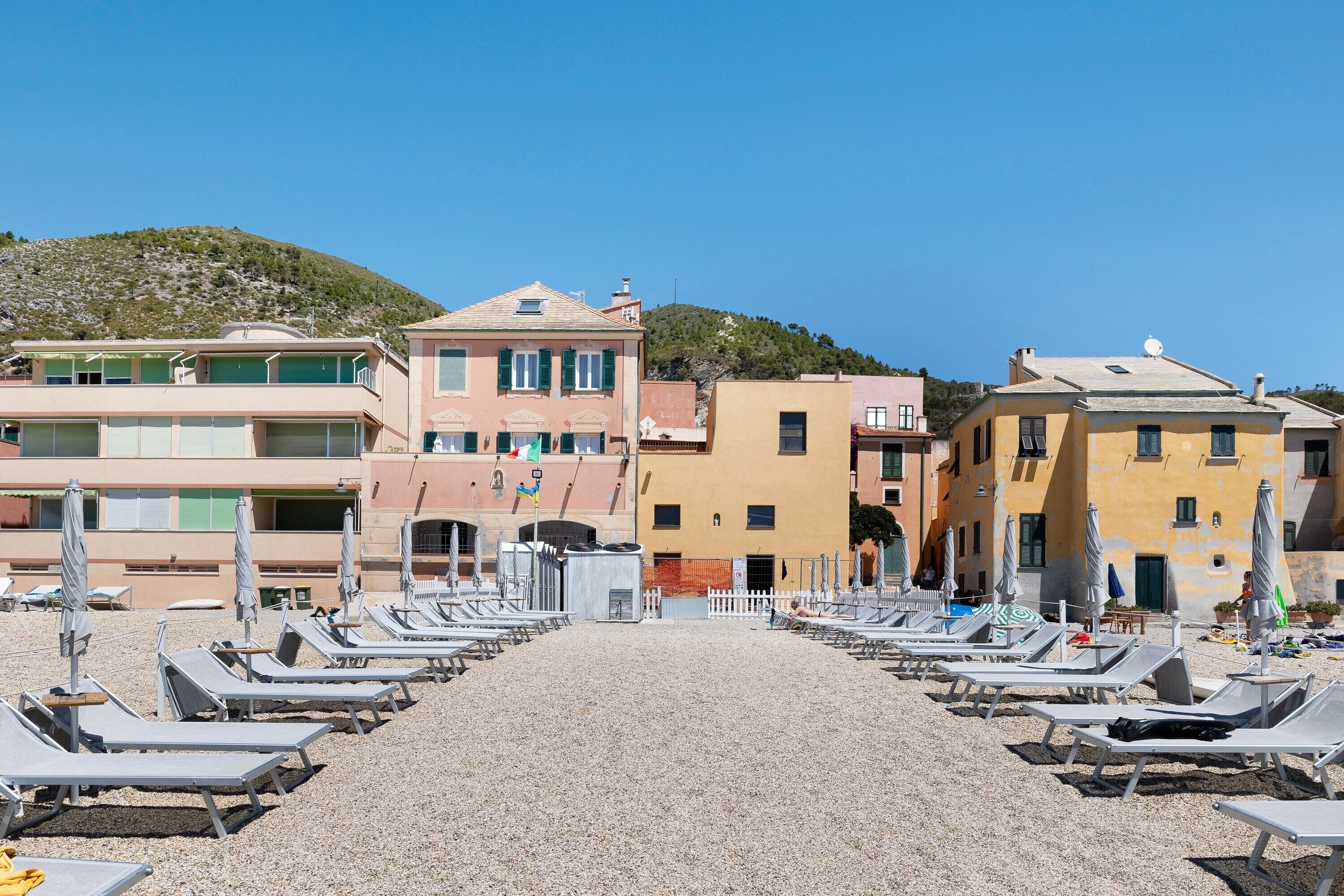 A beachside hotel with rows of sun loungers, a pebble beach, and buildings with balconies.