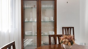 A dining area with a wooden cabinet containing glassware and plates, a round table with a vase of dried flowers, and a wooden chair.