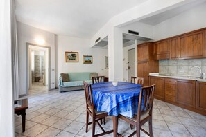 A dining area with a table set for four, wooden cabinets, a kitchen with a sink, and a hallway leading to a bathroom.