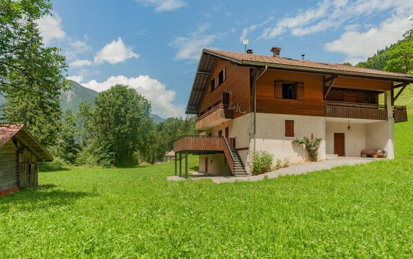 A two-story wooden house with a balcony, a small attached structure, and a grassy hill.