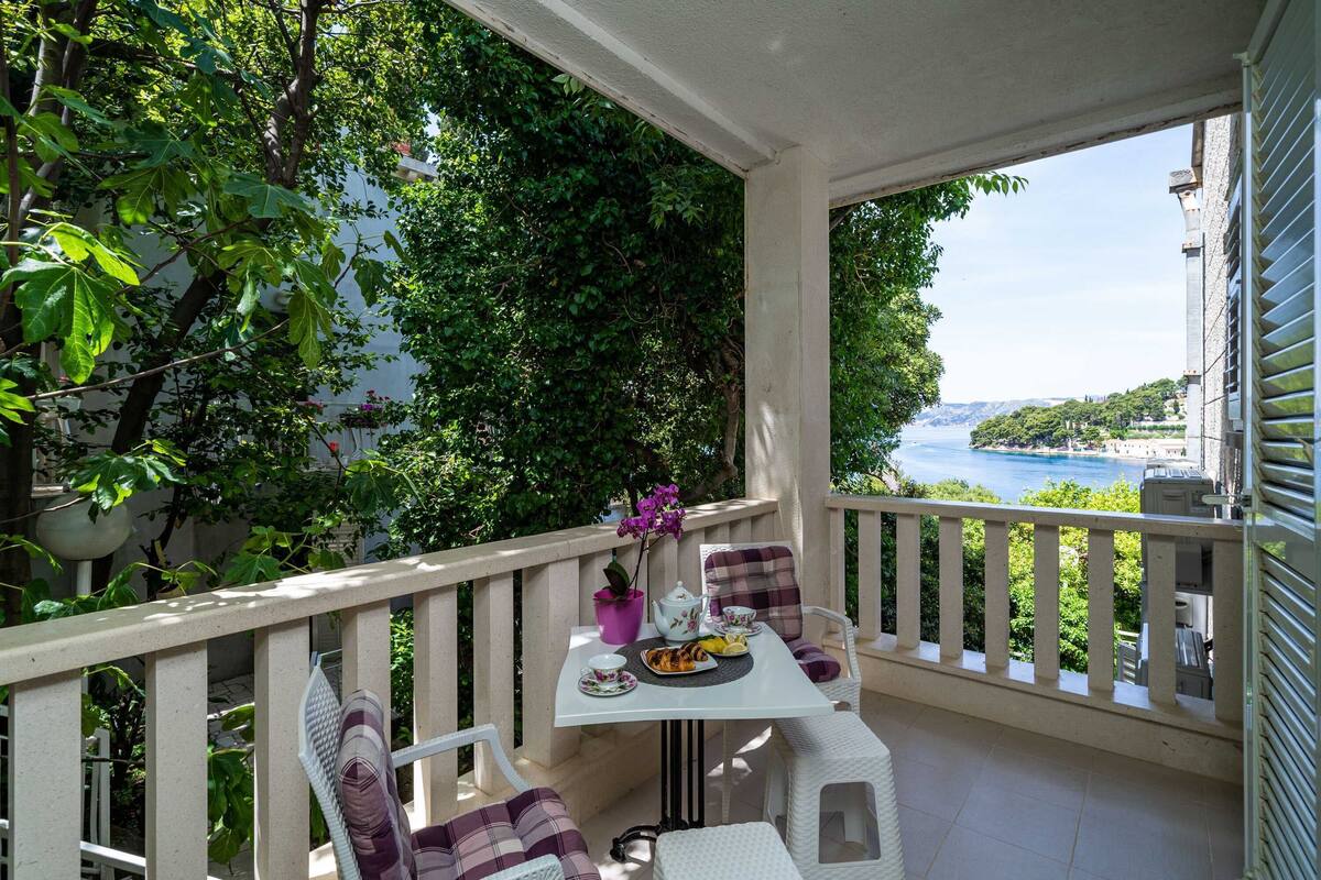 A balcony with a table set for two, surrounded by white wicker chairs, overlooking a scenic view of a coastline.