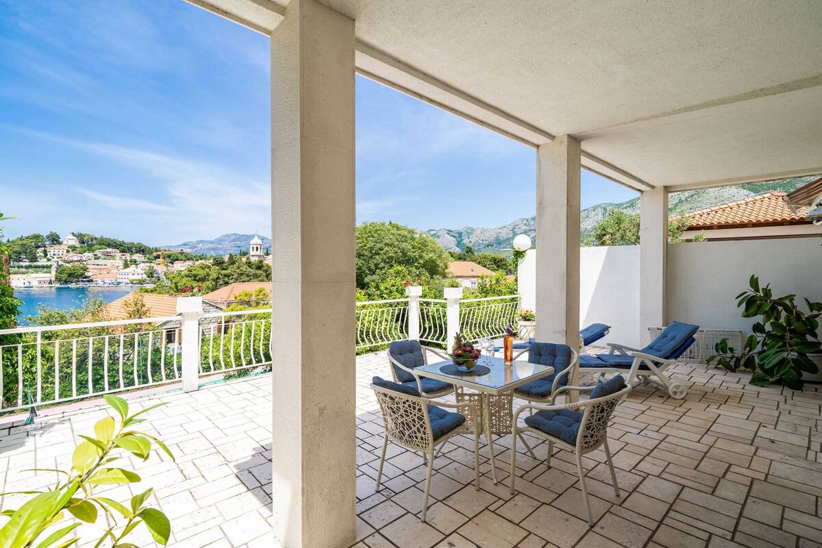 A covered outdoor seating area with a table and chairs, overlooking a scenic view of a coastal town and mountains.
