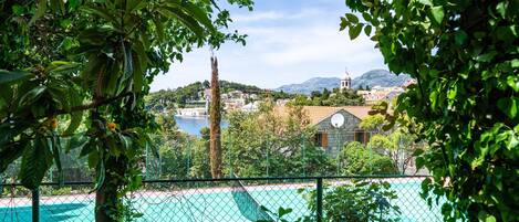 Un campo da tennis circondato da vegetazione rigogliosa, con vista su una città e montagne sullo sfondo.