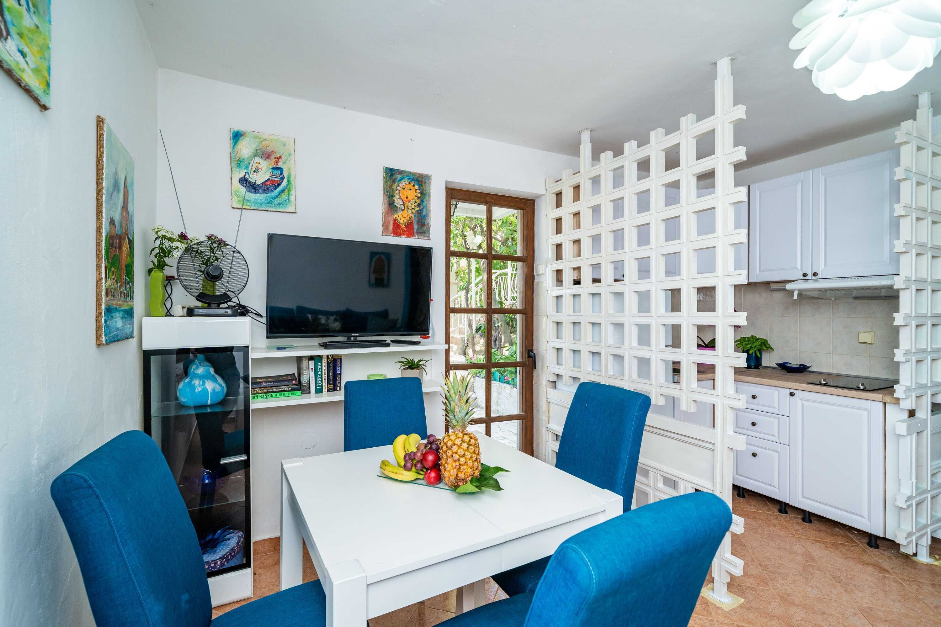 A compact kitchen with white cabinets, a dining area with blue chairs, a TV on a shelf, and artwork on the walls.