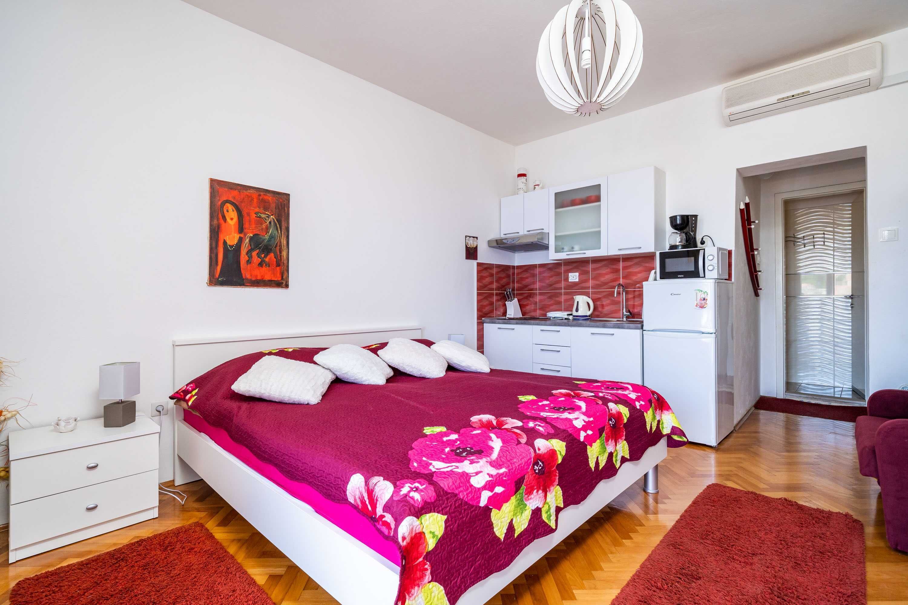 Un dormitorio con cama, mesita de noche, cómoda, alfombra roja, kitchenette con microondas, cafetera y un cuadro en la pared.