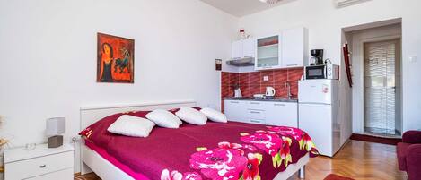 Un dormitorio con cama, mesita de noche, cómoda, alfombra roja, kitchenette con microondas, cafetera y un cuadro en la pared.