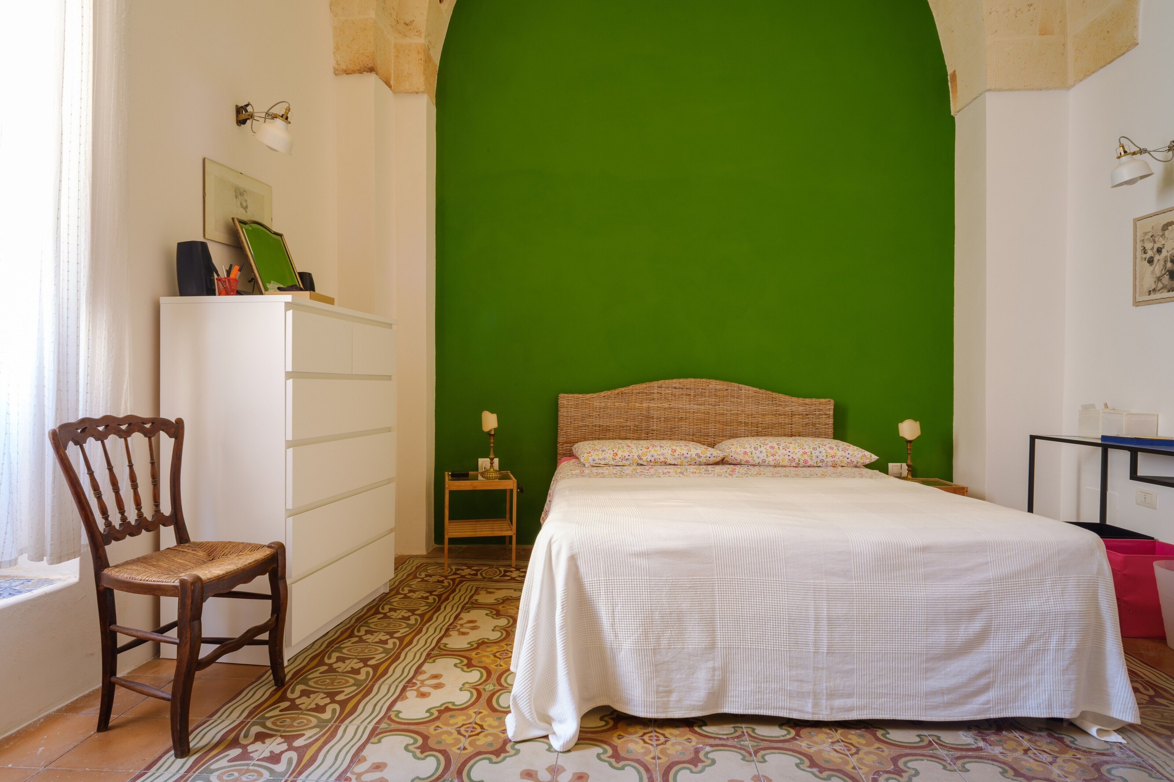 Ein Schlafzimmer mit grüner Wand, einem Bett mit weißer Bettwäsche, einem Holzstuhl und einem gemusterten Teppich.