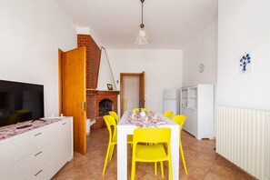 Un coin repas avec une table dressée pour quatre personnes, des chaises jaunes, un meuble blanc, un téléviseur posé sur une commode blanche et une cheminée en briques.