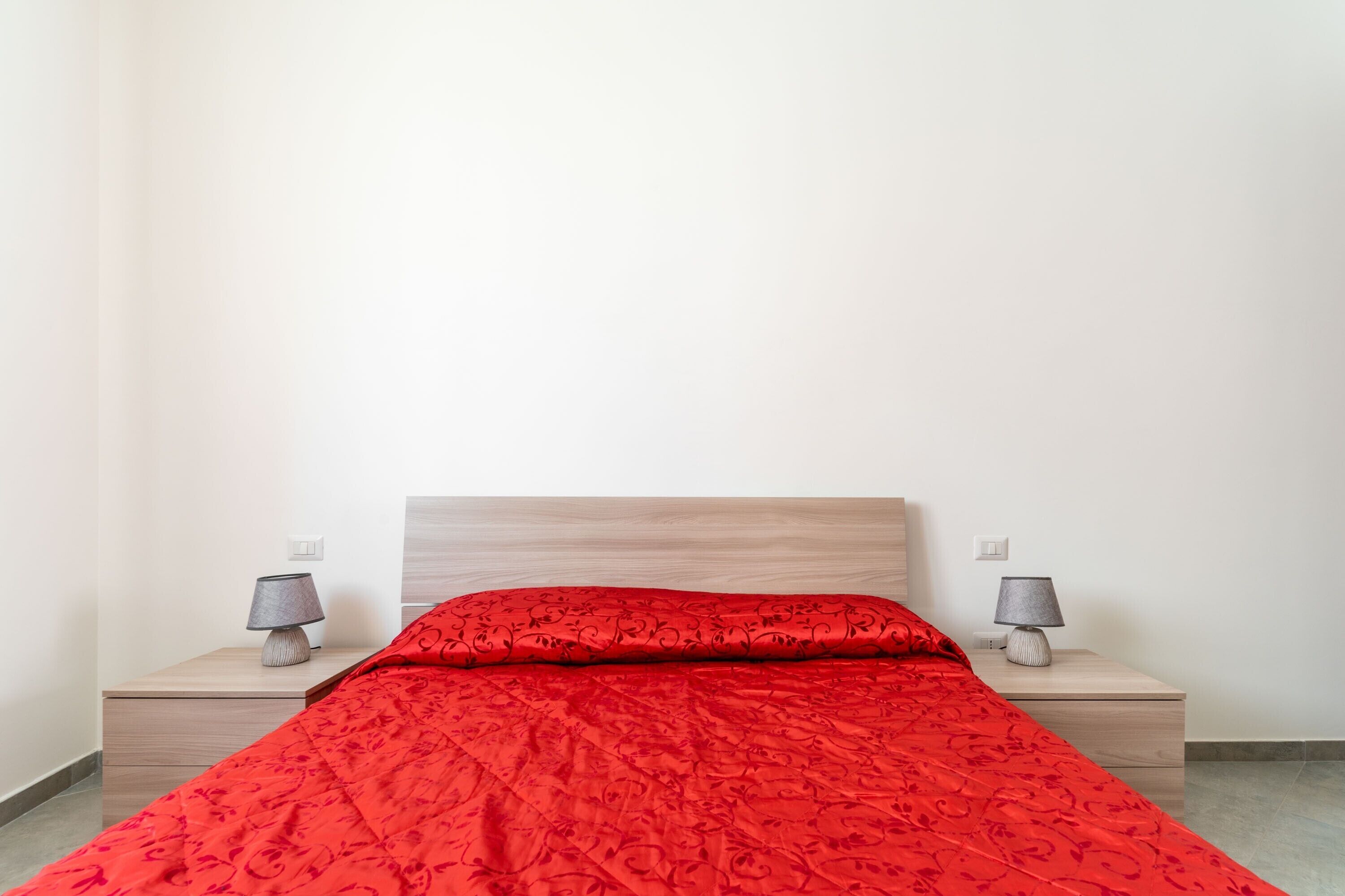 Una cama con una colcha roja estampada, dos lámparas de noche y un cabecero de madera.