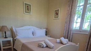 Un lit bien fait, avec du linge de lit blanc et deux serviettes enroulées, une table de chevet avec une lampe et une fenêtre avec des rideaux.