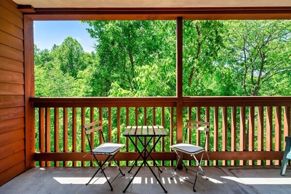 Un balcón de madera con una mesa y dos sillas, con vista a un bosque verde y frondoso.