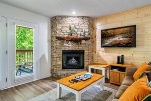 Una sala de estar con chimenea de piedra, un televisor montado en la pared y vista a zonas verdes a través de una ventana.