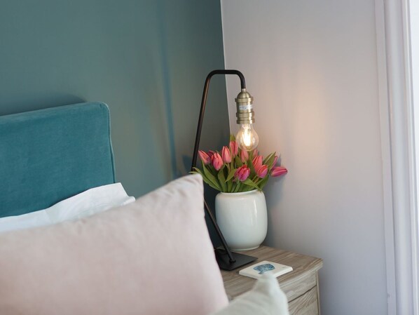 A neatly made bed with teal and white bedding, a white vase with pink flowers, and a bedside lamp on a wooden nightstand. - Marchbourne (St Ives)