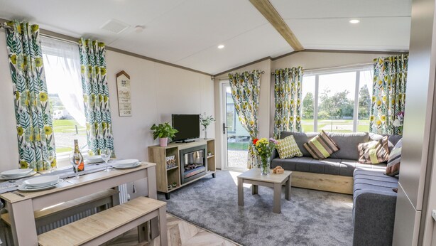 A modern living room with a sofa, coffee table, and TV stand. There is a dining table with chairs and a bench. The room has large windows with curtains. - Jean's Place (Malton)
