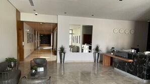 A modern reception area with a marble reception desk, two potted plants, a seating area with chairs, and a hallway leading to a room with a patterned wall. - VOYAGE RESIDENCE (Riyadh)