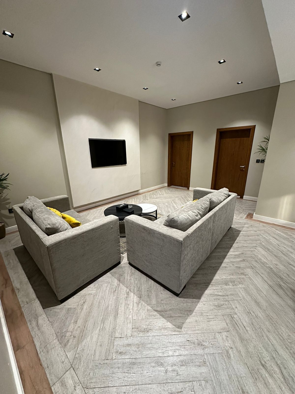 A spacious living area with two grey sofas, a flat-screen TV mounted on the wall, and a herringbone-patterned floor.