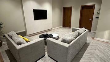 A spacious living area with two grey sofas, a flat-screen TV mounted on the wall, and a herringbone-patterned floor.