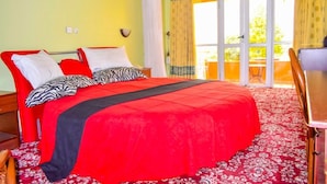 A room with a large bed, a wooden desk, and a balcony with yellow curtains.