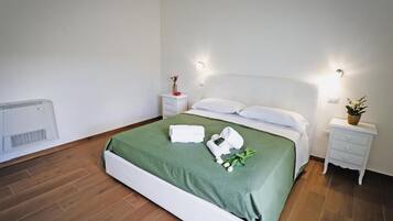Un lit bien fait, recouvert d’une couvre-lit verte, avec des oreillers blancs et une tête de lit blanche. Il y a deux tables de chevet, chacune munie d’une lampe, placées de chaque côté du lit. Une petite plante en pot se trouve sur la table de chevet à droite.