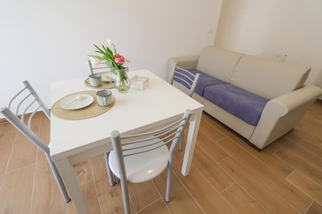 Un coin repas avec une table blanche, quatre chaises, un vase de fleurs et un canapé beige agrémenté d’un coussin violet.