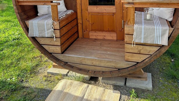 Una habitación con forma de barril de madera, con una puerta, dos zonas de estar acolchadas y una pequeña mesa con una vela.