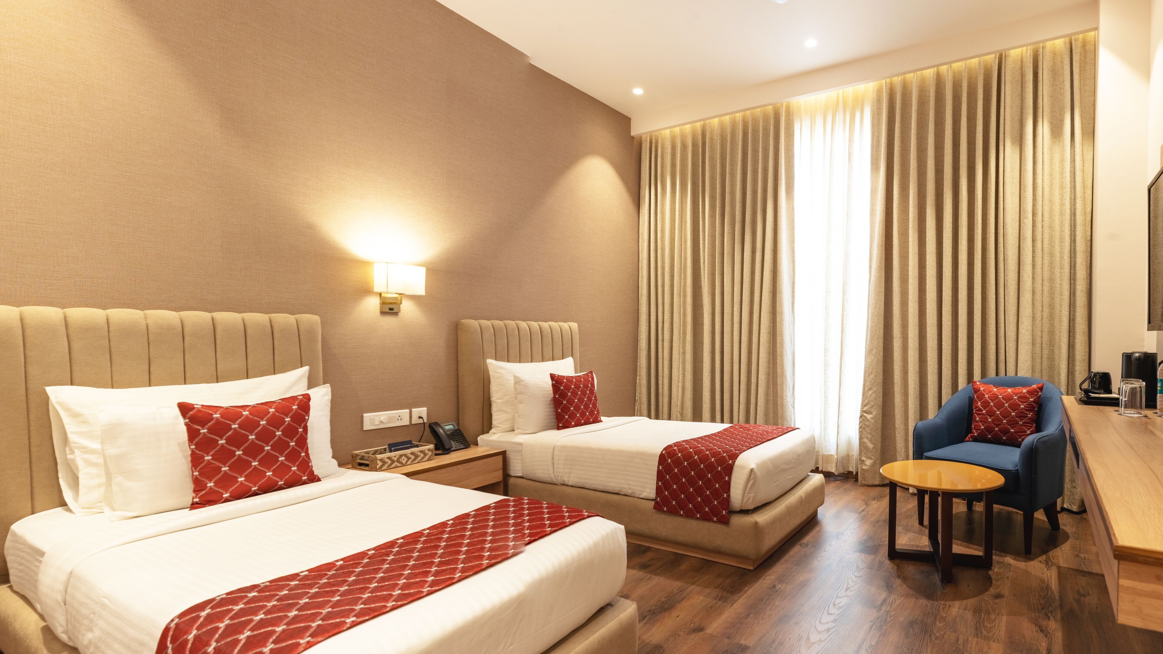 A hotel room with two beds, a desk, a chair, a lamp, and a large window with curtains.