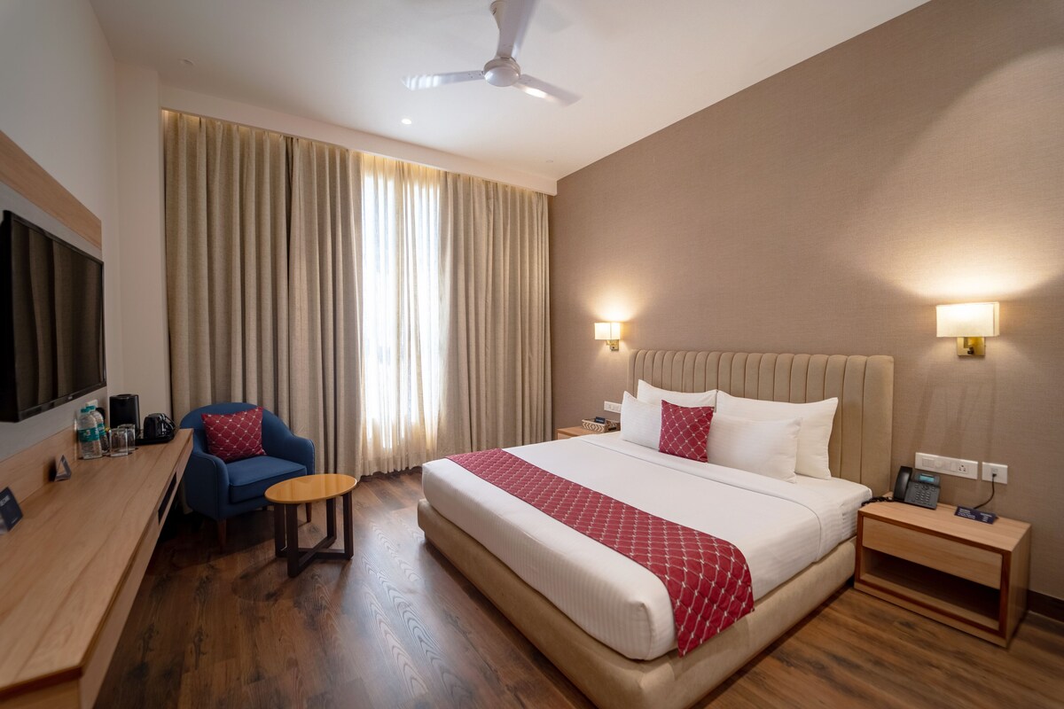 A hotel room with a large bed, a desk with a chair, a small table, a TV, and a ceiling fan.