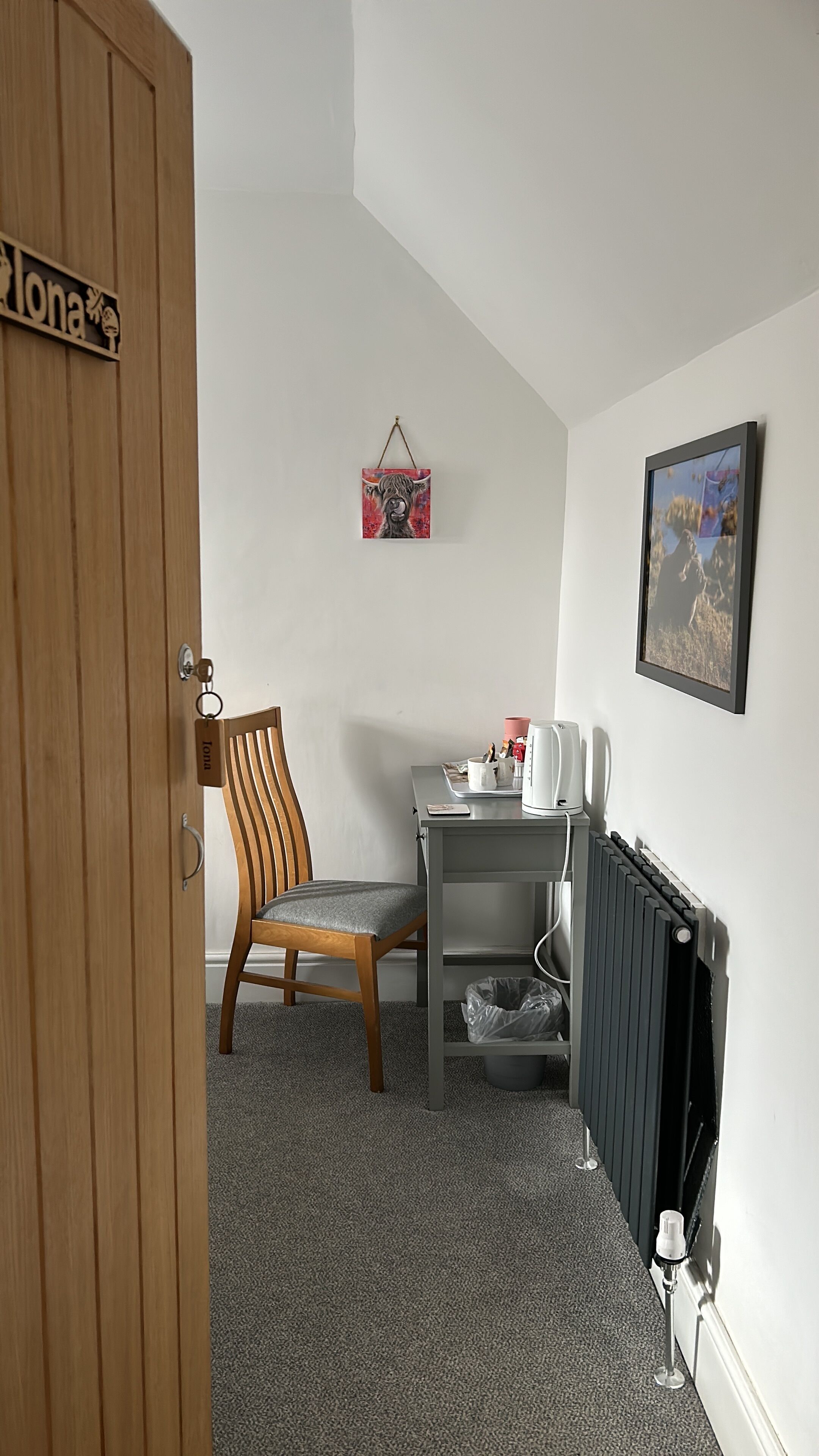 Una habitación con puerta de madera, una silla, una mesita con una tetera, un radiador y un cuadro en la pared.