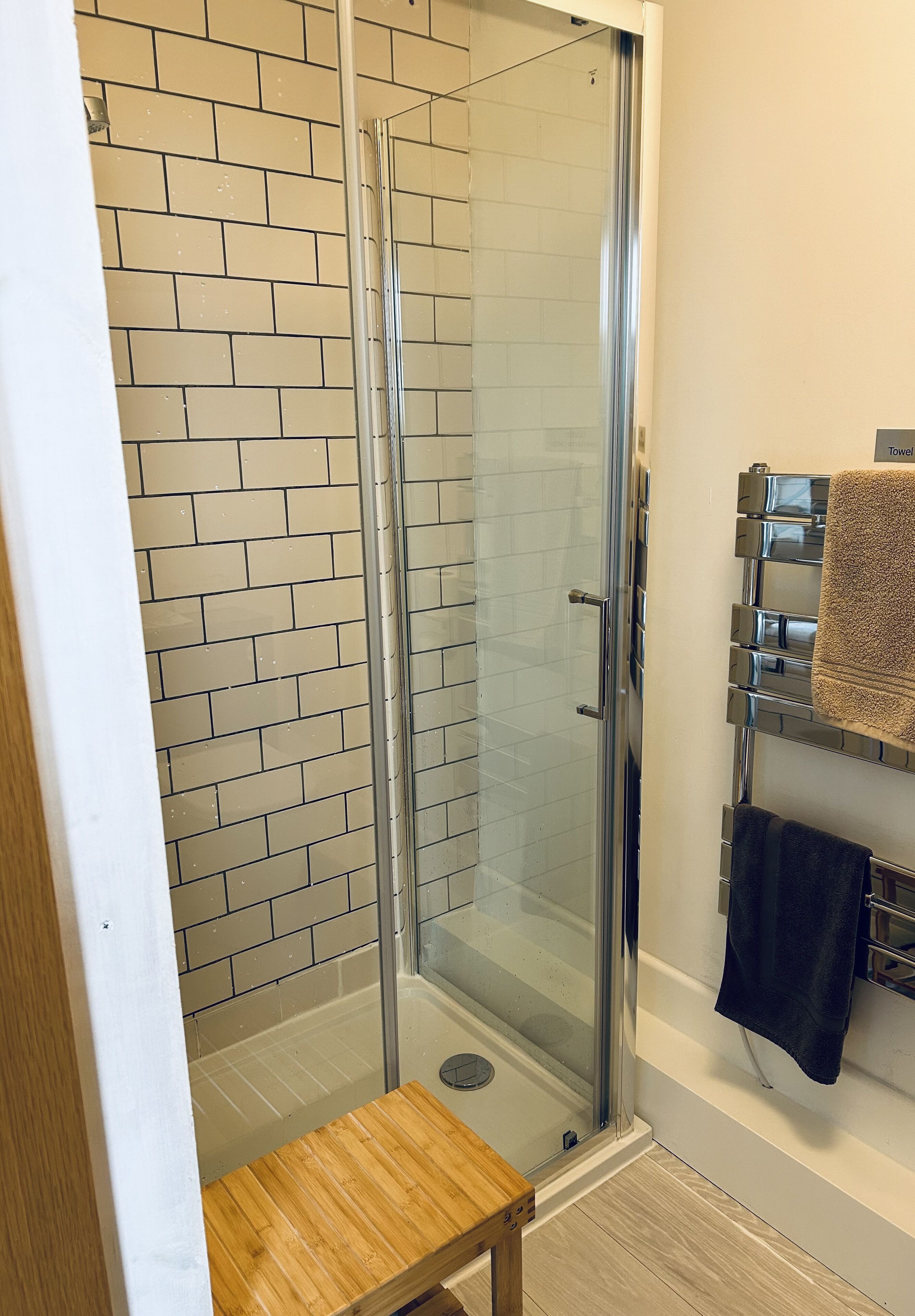 Ducha con paredes de vidrio, pared de azulejos, banco de madera y toallero con toallas.