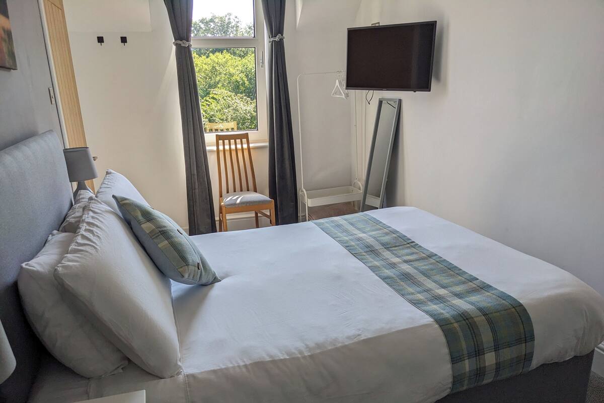 Una cama bien hecha con una manta escocesa, una silla de madera y un televisor montado en la pared.