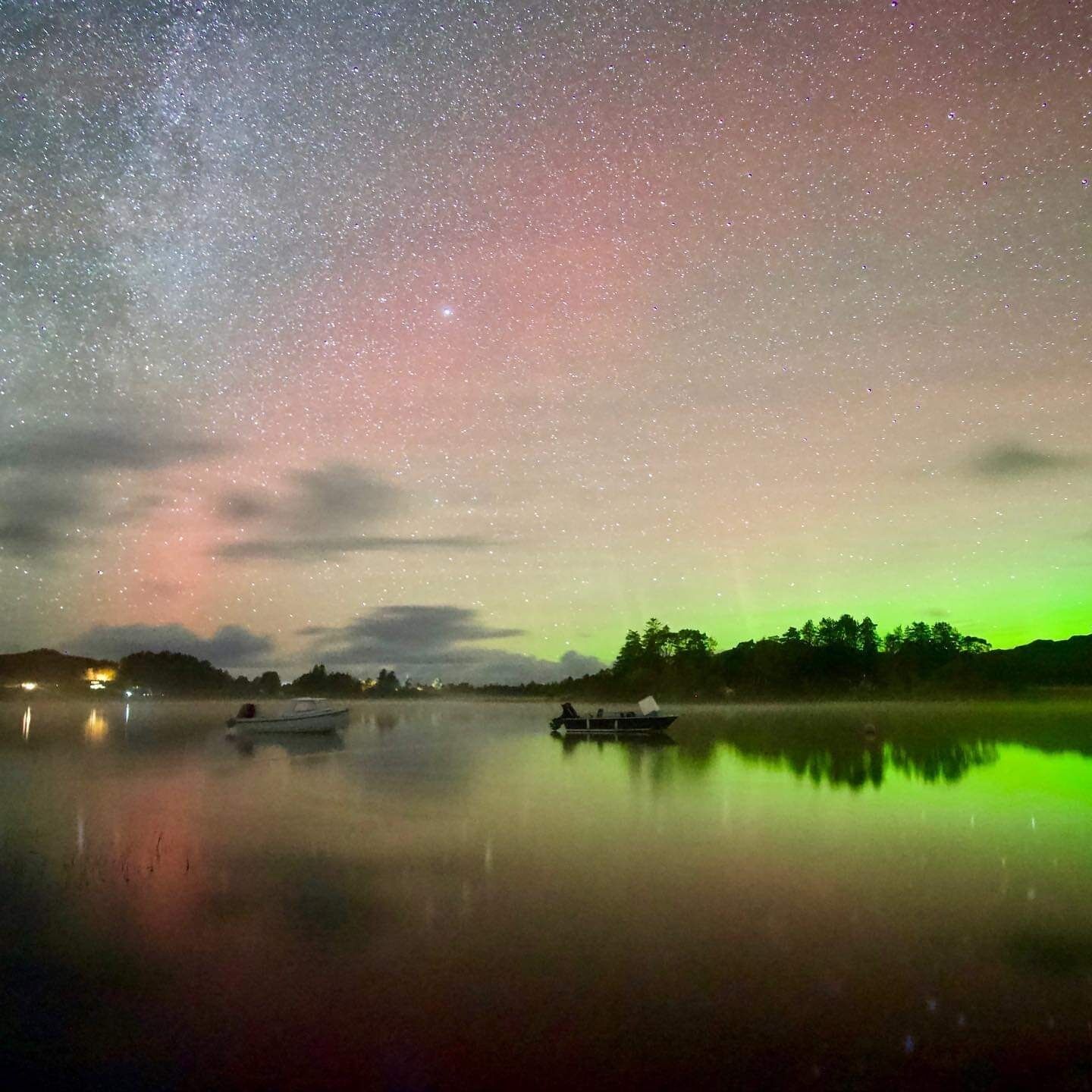 A night sky with stars and the aurora borealis over a calm lake with boats.