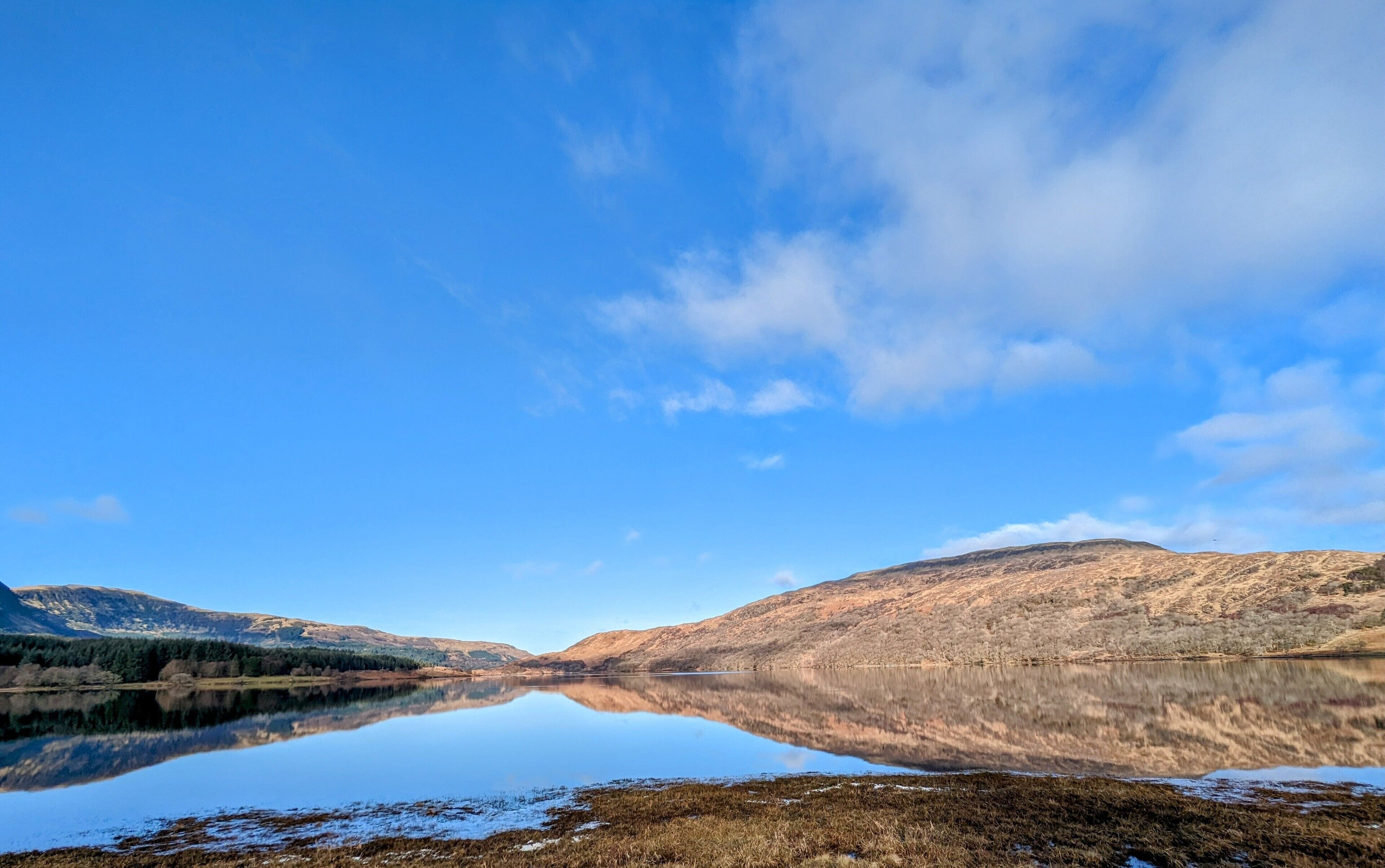 A calm lake with clear reflections, surrounded by hills and sparse vegetation.