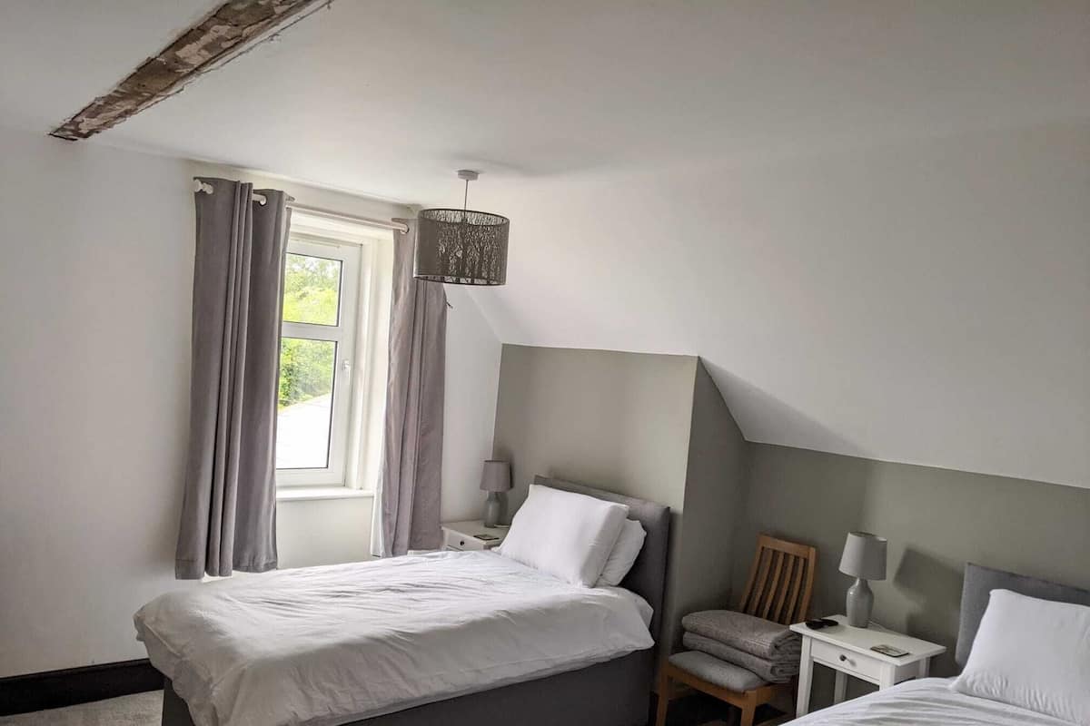 Un dormitorio con dos camas, una ventana con cortinas, una mesita de noche con lámpara y una silla.