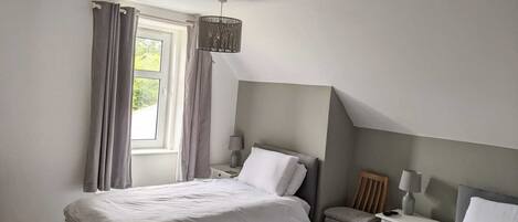 A bedroom with two beds, a window with curtains, a bedside table with a lamp, and a chair.