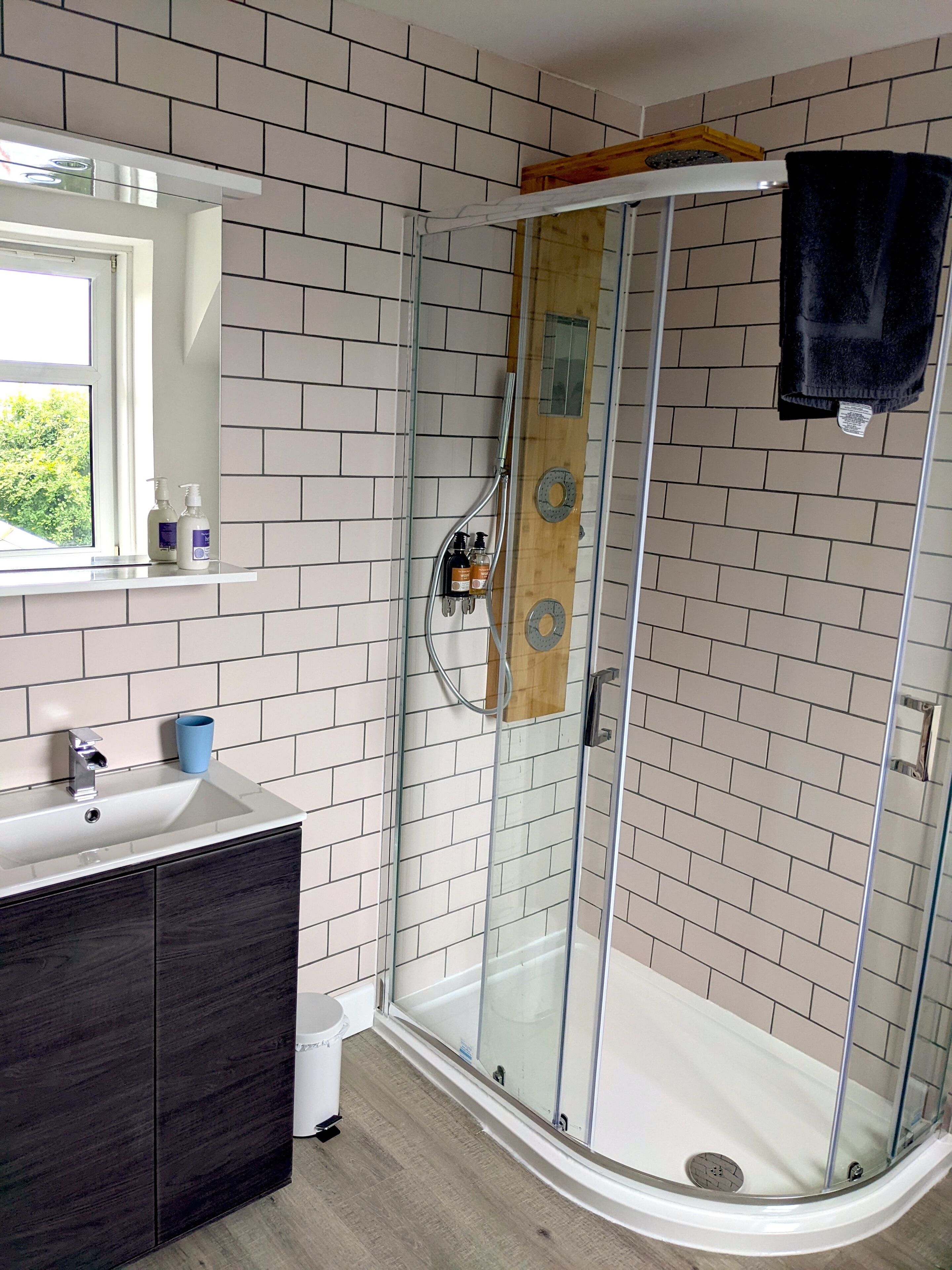 Un baño moderno con ducha envuelta en vidrio, un lavamanos blanco, armarios de madera oscura y una ventana con vista a la vegetación.