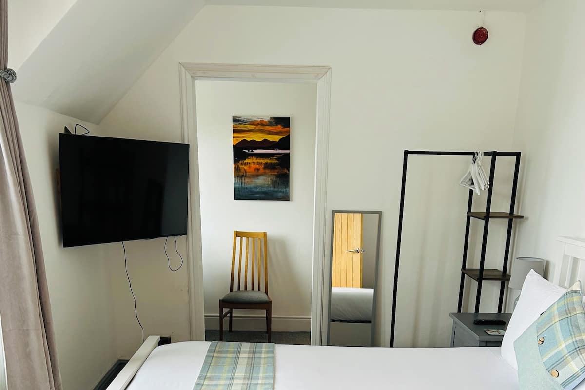 Una cama bien hecha con una manta escocesa, un televisor montado en la pared, una silla y una puerta que da a otra habitación.