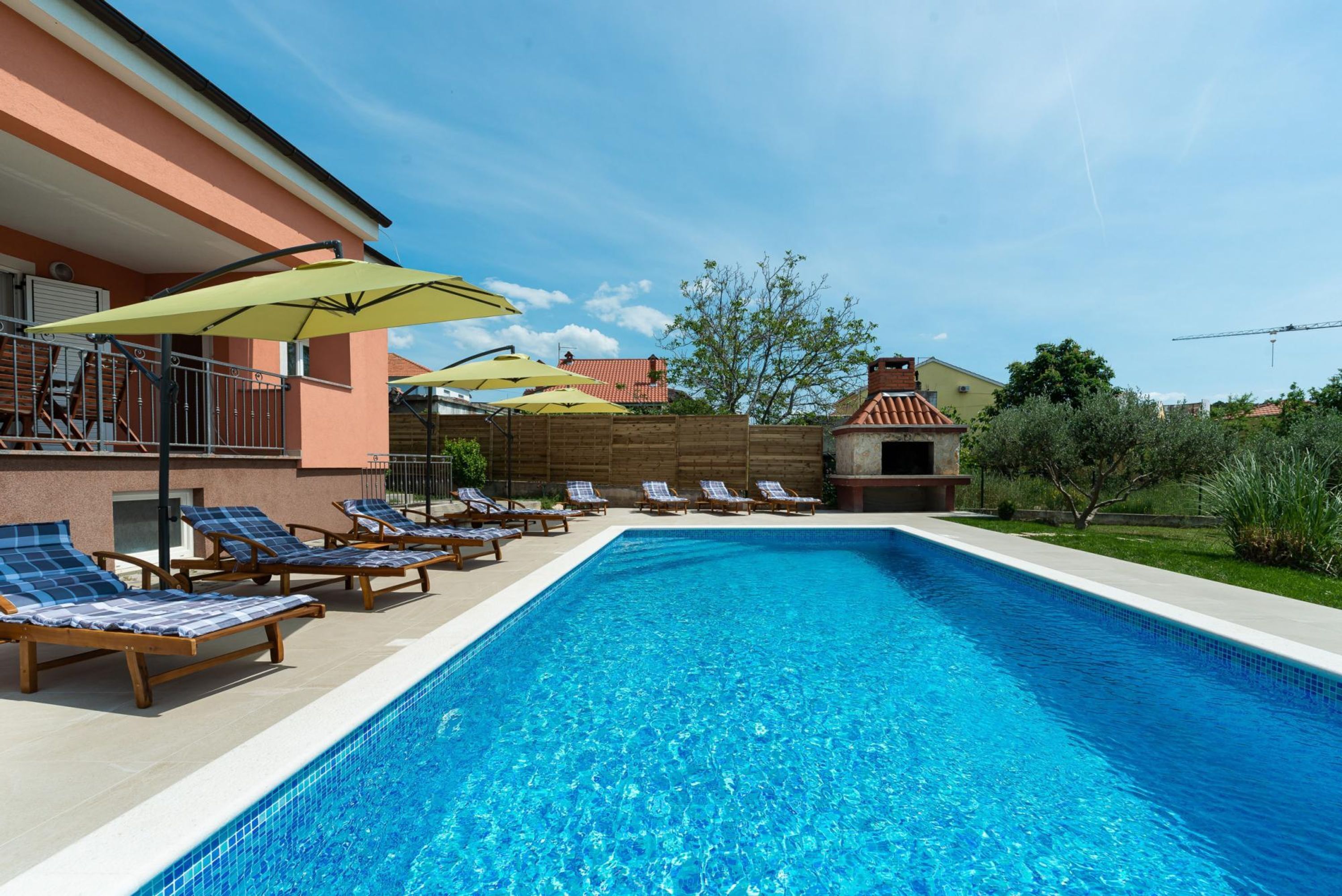 Una piscina con sillones de descanso y sombrillas, rodeada por un edificio con techo de tejas y pared de ladrillo.