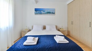 Ein ordentlich bezogenes Bett mit einer blauen Bettdecke, weißen Kissen und zwei aufgerollten Handtüchern. Darüber an der Wand hängt ein Gemälde einer Welle.