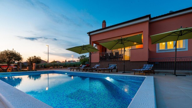 Ein Schwimmbecken mit Liegestühlen und Sonnenschirmen, ein Gebäude mit Balkon und ein klarer Himmel.