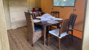 A wooden dining area with a table set for four, surrounded by wicker chairs. A window with a view of trees is visible. - Glamping TENTE LODGE SAFARI DE LUXE, vue sur le coteau et vignes (Chadenac)