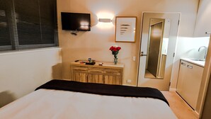 Et hotelværelse med en seng, et natbord med en vase blomster, et fjernsyn ophængt på væggen og et badeværelse med et spejl og en vask.