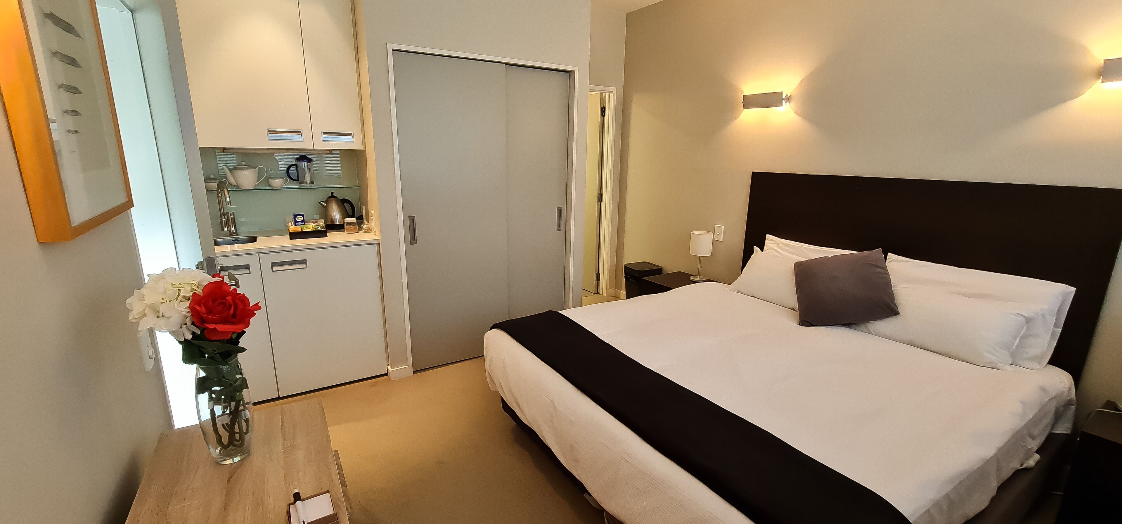 A hotel room with a bed, bedside table, nightstand, and a small kitchenette area.