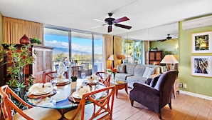 A spacious living area with a dining table set for four, surrounded by wicker chairs. A sofa and armchairs are arranged in the living room, with a large window offering a view of the outdoors.