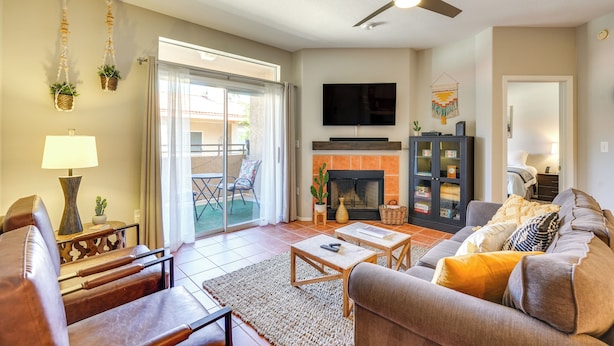 A living room with a sofa, coffee table, and TV. There is a fireplace, a ceiling fan, and a sliding glass door leading to an outdoor area.