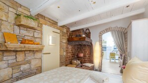 A cozy room with a stone wall, a bed with a patterned bedspread, a small shelf with decorative items, and a view of the outside through an arched doorway. - Apartment Alba (Kastelir-Labinci)