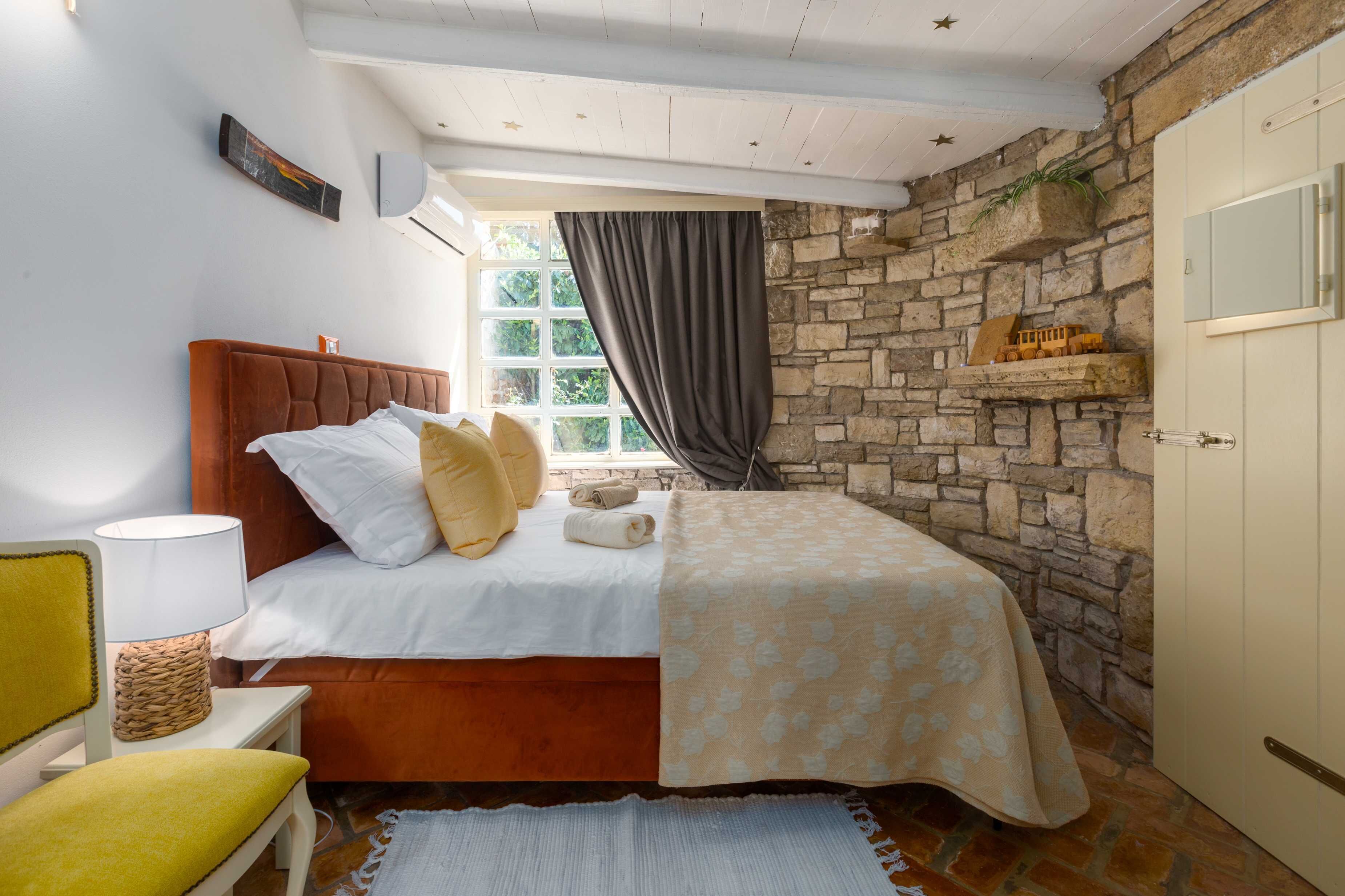 A bedroom with a stone wall, a bed with a wooden headboard, a bedside table with a lamp, and a window with curtains.