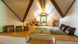 Een knus slaapkamer onder een schuine kap, met houten vloeren, een groen vloerkleed, een bank met kussens, een eettafel met stoelen en een raam met gordijnen.