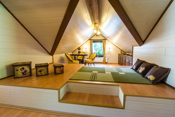 Ein gemütliches Schlafzimmer im Dachgeschoss mit schrägen Wänden, Holzboden, einem grünen Teppich, einer Couch mit Kissen, einem Esstisch mit Stühlen und einem Fenster mit Vorhängen.