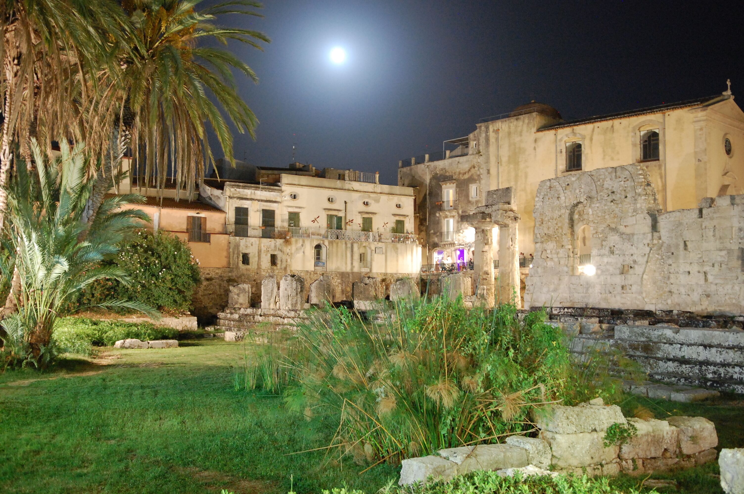 An ancient stone building with a courtyard, palm trees, and a full moon in the background.